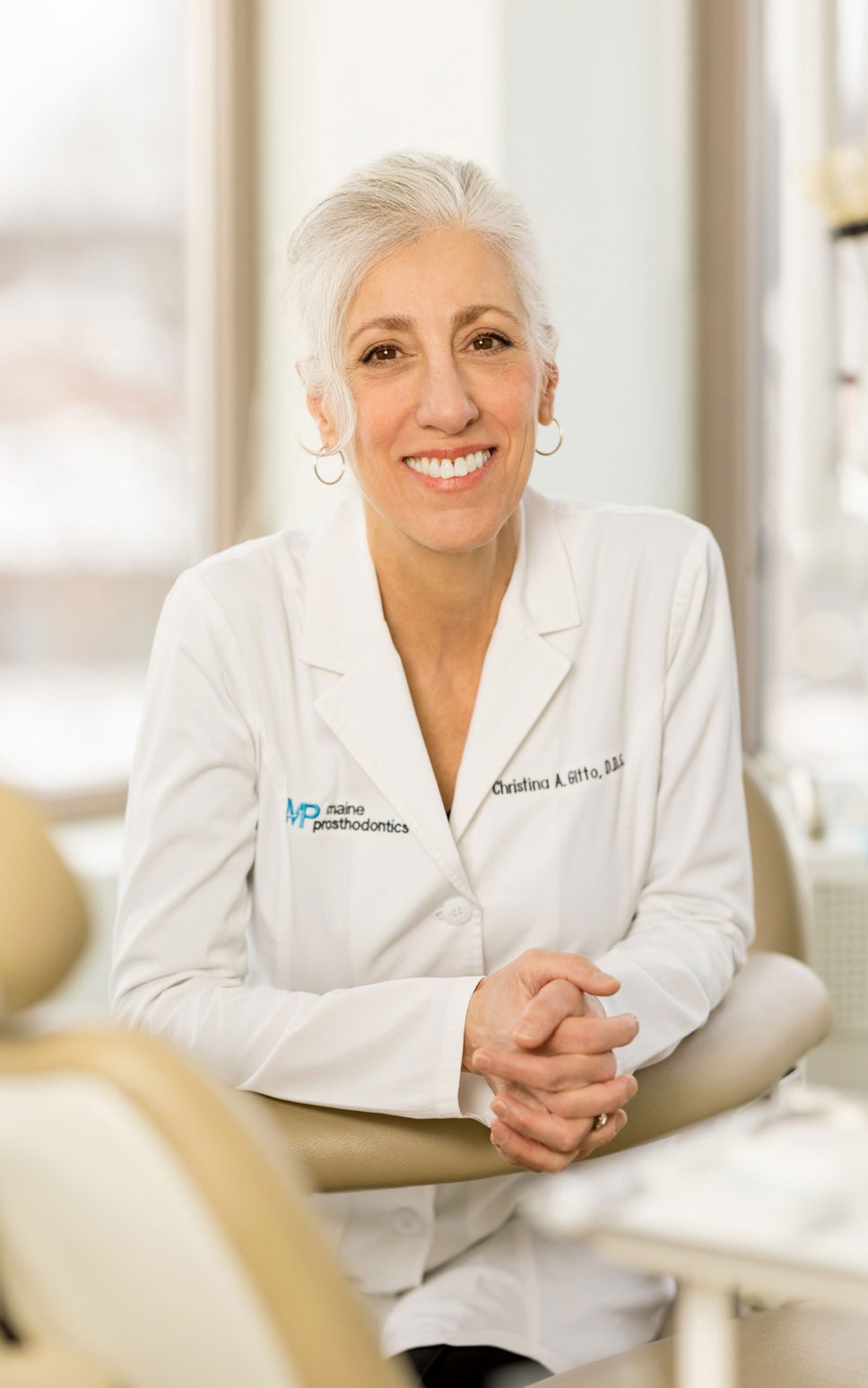 A woman in a white lab coat leans against her desk and smiles.