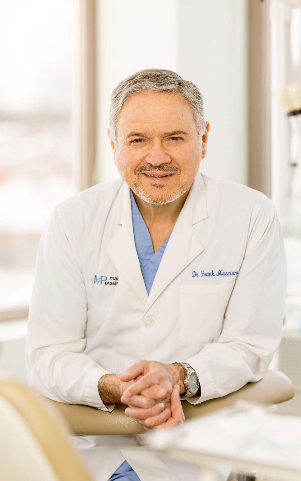 A man in a white lab coat and medical scrubs smiles with hands folded.